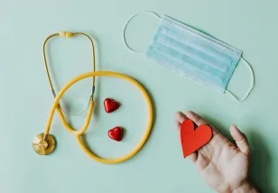 A yellow stethoscope, blue paper face mask, and a hand holding a red felt cut-out heart