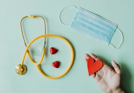 A yellow stethoscope, blue paper face mask, and a hand holding a red felt cut-out heart